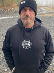 Man wearing a black hoodie with a logo and text, standing outdoors on a gravel surface.
